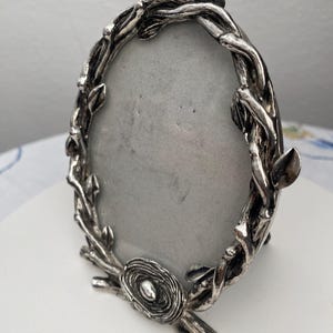 Puede incluir: Marco de fotos ovalado plateado con un diseño decorativo de ramitas y hojas. El marco tiene un pequeño detalle de nido en la parte inferior. El marco está vacío, con un interior gris.