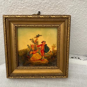 Puede incluir: Pequeña obra de arte enmarcada cuadrada con un marco dorado y ornamentado. La obra representa a una pareja con ropa de época, una mujer tocando un laúd y un hombre con un abrigo rojo. El fondo es un paisaje con árboles y un cielo amarillo.