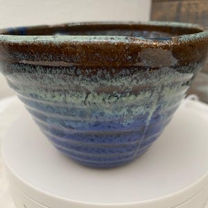 Może przedstawiać: Ręcznie robiona ceramiczna miska o teksturowanej powierzchni. Miska ma gradient kolorów, w tym brązowy, niebieski i turkusowy. Miska ma szeroki brzeg i zaokrąglony kształt, idealna do serwowania jedzenia lub jako element dekoracyjny.