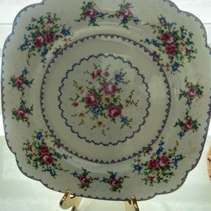 May include: A square, white plate with a scalloped edge and a gold rim. The plate features a cross-stitch floral design in pink, blue, green, and yellow. The plate is displayed on a gold-colored stand.