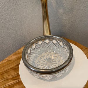 Op de afbeelding: Een kom van helder glas met een diamantgeslepen patroon aan de buitenkant en een sterontwerp op de bodem. De kom is afgewerkt met een gladde, gepolijste zilverkleurige metalen rand aan de bovenkant, wat zorgt voor een strakke afwerking.