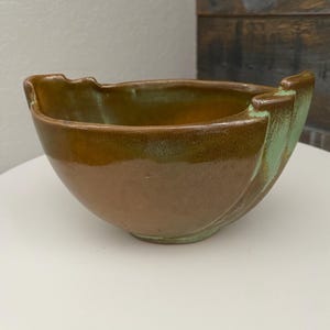 Puede incluir: Un cuenco de cerámica hecho a mano con un diseño único. El cuenco presenta un exterior marrón con un borde y detalles verdes. El cuenco tiene una forma curva y probablemente se utiliza para servir o exhibir.