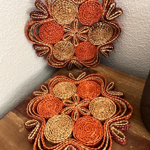 Puede incluir: Dos posavasos decorativos redondos hechos de fibras naturales tejidas. Los posavasos presentan un diseño floral con tonos naranja, beige y burdeos. Los posavasos están colocados sobre una superficie de madera y contra una pared blanca.