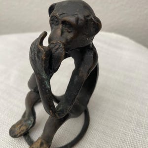 May include: A bronze monkey figurine in a seated pose, with its hand raised to its mouth. The monkey's tail forms a circular base. The figurine has a detailed face and is made of a dark, metallic material.