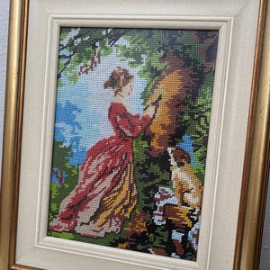 May include: Framed needlepoint art depicting a woman in a red dress and a dog in a forest setting. The artwork is framed in a gold-colored frame with a cream-colored mat. The scene includes trees, blue sky, and detailed stitching.