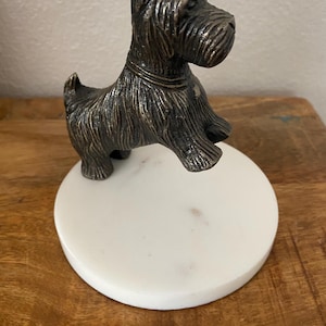 May include: A bronze-colored Scottie dog figurine atop a white marble coaster. The dog has detailed fur and a collar. The coaster is round and sits on a wooden surface. A decorative item.