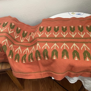 May include: Rust-colored textile with a repeating pattern of green and cream-colored tulip-like floral designs. The fabric is draped over a wooden surface, showcasing the detailed embroidery and the scalloped edge of the textile.