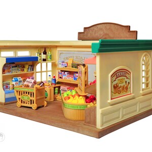 May include: A miniature toy grocery store with shelves stocked with food items, a shopping cart, and a bakery section. The store has a tan exterior with green trim and a brown roof. The bakery sign features illustrations of bread and pastries.