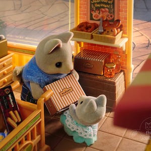 May include: A miniature bakery scene with animal figurines. A gray cat figurine looks over a shelf of snacks. Two bear figurines hold a bakery box. The bakery shelf displays baked goods and a sign that reads "Just out of the oven!"