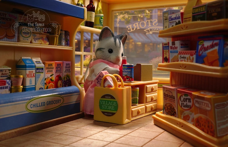 May include: A miniature grocery store scene with a cat figurine wearing a pink apron. The store features shelves stocked with various food items, including juice boxes, milk cartons, and canned goods. A yellow shopping bag with "VILLAGE STORE" text is in the foreground.
