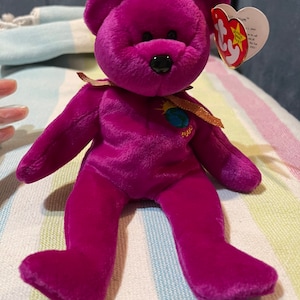 Puede incluir: Oso de peluche morado Beanie Baby con una cinta marrón y una etiqueta que dice "Beanie Babies" y "2000".