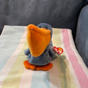 May include: A blue and orange plush pelican with a red Ty tag that says "Ty" and has a star on it. The pelican is sitting on a striped towel.
