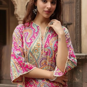 May include: A woman wearing a pink paisley print tunic with a V-neckline and three-quarter sleeves. The tunic has white trim and a colorful paisley pattern in shades of pink, blue, yellow, and purple. The sleeves are flared.