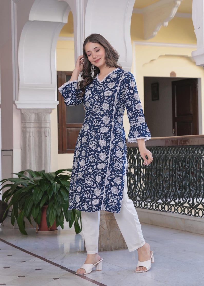 May include: A long-sleeved, navy blue tunic with a white floral pattern and white lace trim. The tunic is paired with white pants and white heeled sandals. The outfit is suitable for casual or semi-formal wear.