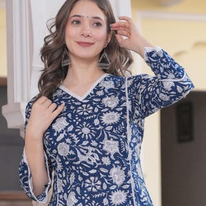 May include: A long-sleeved, navy blue tunic with a white floral pattern. The tunic has a V-neckline and white lace trim. The sleeves are three-quarter length. The model is wearing triangle-shaped earrings.