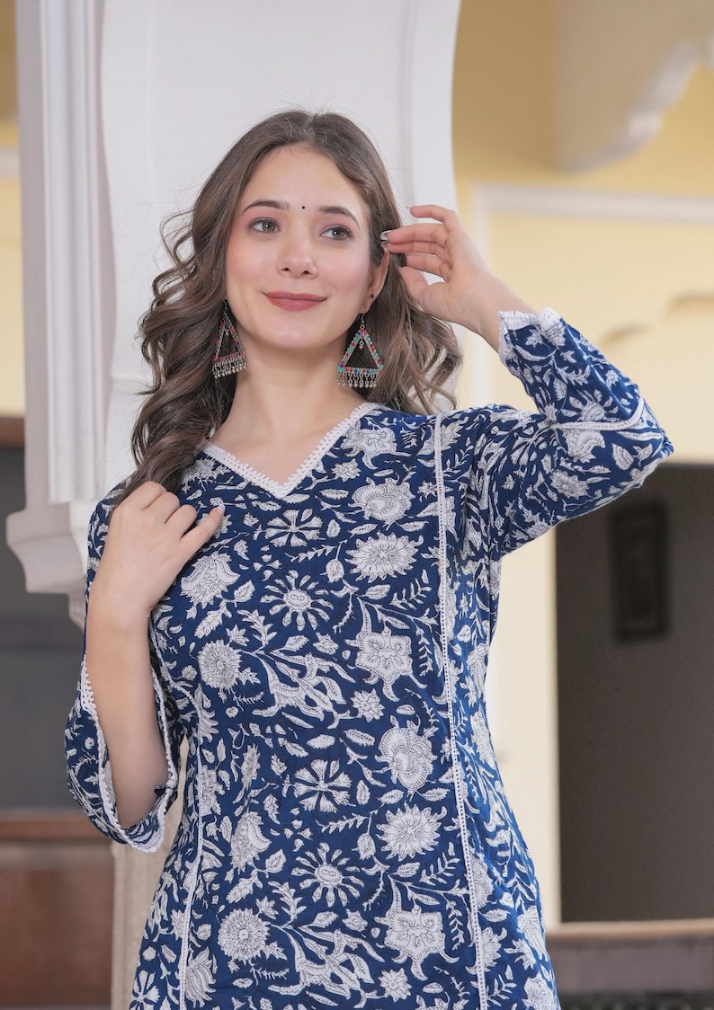 May include: A long-sleeved, navy blue tunic with a white floral pattern. The tunic has a V-neckline and white lace trim. The sleeves are three-quarter length. The model is wearing triangle-shaped earrings.