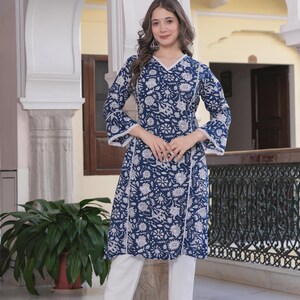 May include: A woman wearing a blue floral print tunic with white pants. The tunic has a V-neck and white lace trim. The outfit is a traditional style. The background includes architectural details and a potted plant.