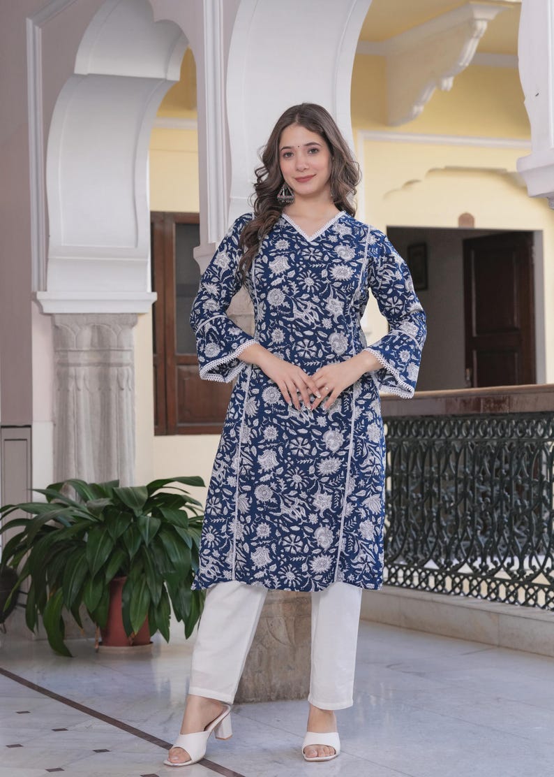 May include: A woman wearing a blue floral print tunic with white pants. The tunic has a V-neck and white lace trim. The outfit is a traditional style. The background includes architectural details and a potted plant.