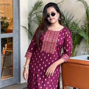 Puede incluir: Un conjunto kurta de color ciruela con cuello cuadrado y mangas tres cuartos. El kurta presenta un estampado de lunares dorados y bordados intrincados en rojo y dorado. Las mangas y el escote están adornados con patrones detallados.