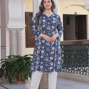 May include: A woman wearing a navy blue tunic and white pants. The tunic features a floral pattern in white and a white trim. The pants are straight-legged. The woman is wearing white heels.