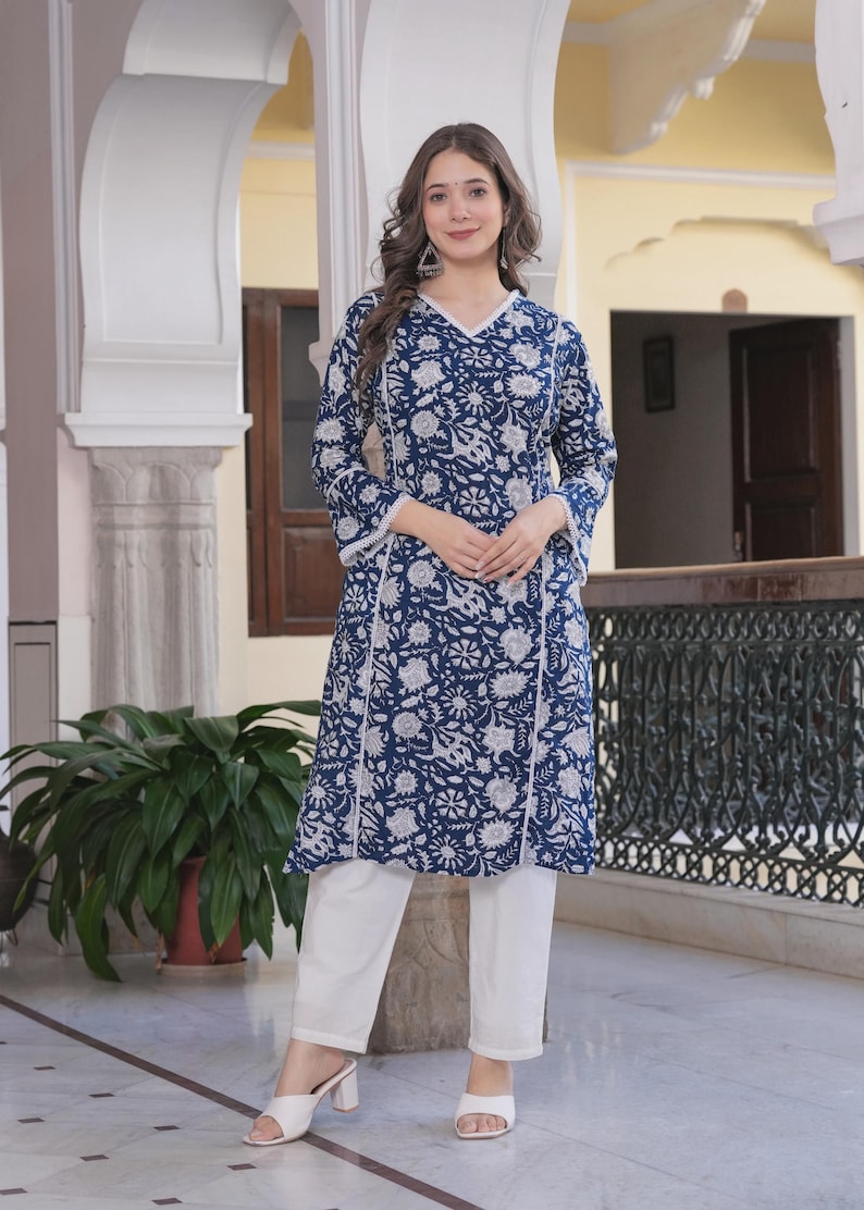 May include: A woman wearing a navy blue tunic and white pants. The tunic features a floral pattern in white and a white trim. The pants are straight-legged. The woman is wearing white heels.