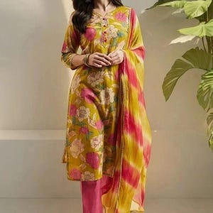 May include: A mustard yellow floral print salwar kameez set. The outfit includes a long-sleeved tunic, matching pants, and a dupatta. The tunic and dupatta feature a pink and white floral pattern. The dupatta has pink and yellow stripes.