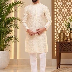 Pode incluir: Um kurta de cor creme de manga comprida com um padrão floral tonal subtil, gola mandarim e uma carcela de botões parcial, combinado com calças brancas simples e sapatos castanhos sem atacadores. A peça tem uma bainha reta.