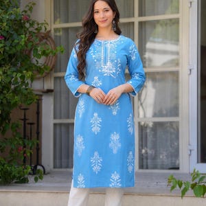 Peut inclure: Ensemble tunique bleu clair et pantalon blanc. La tunique présente des broderies florales blanches sur l'encolure, les manches et le devant. La tenue est complétée par un pantalon blanc et des talons dorés. Le modèle est sur des marches blanches.