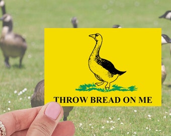 May include: A yellow sign with a black and white illustration of a goose. The sign reads "Throw Bread On Me".