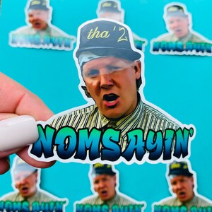 May include: A sticker featuring a man wearing a backwards baseball cap with the text "tha' 2" on it. The man is yelling and the sticker has the text "NOM SAYIN'" in a colorful, graffiti-style font.
