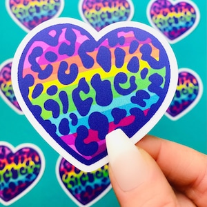 May include: A rainbow heart-shaped sticker with a leopard print design. The sticker has a white border and is made of vinyl.
