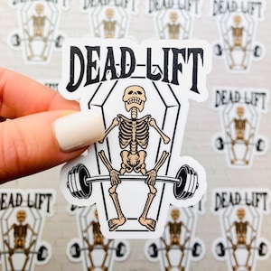 May include: A white sticker with a black and white illustration of a skeleton lifting a barbell inside a coffin. The text "DEAD-LIFT" is written in black at the top of the sticker.