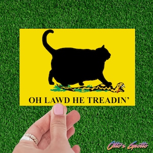May include: A yellow flag with a black silhouette of a cat stepping on a green snake. The text "OH LAWD HE TREADIN'" is printed below the image.