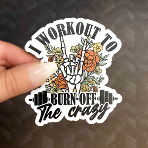 May include: A white sticker with black text that reads "I workout to burn off the crazy". The sticker features a skeleton hand making the rock and roll hand gesture with flowers behind it.