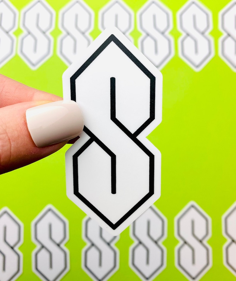 May include: A white sticker with a black outline of the letter 'S' in a stylized, geometric font. The sticker is being held by a hand against a bright green background.