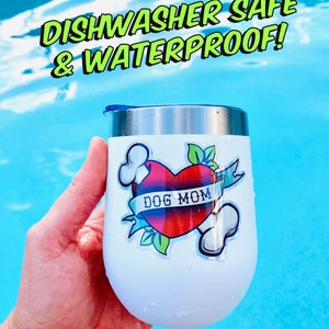 May include: White stainless steel tumbler with a blue lid and a sticker that says "Dog Mom" with a red heart and bones. The tumbler is in a hand and the background is a blue pool.