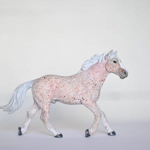 May include: A toy horse figurine in a walking pose. The horse is primarily white with red speckles and a white mane and tail. The hooves are black. The horse is set against a plain white background.