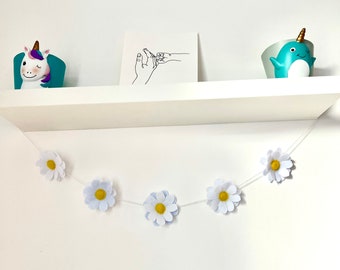 Puede incluir: Un estante flotante blanco sostiene una maceta de unicornio, una de narval y una tarjeta cuadrada con un dibujo lineal negro de una mano tocando una pata. Una guirnalda de cinco margaritas de fieltro blanco con centros amarillos cuelga debajo.