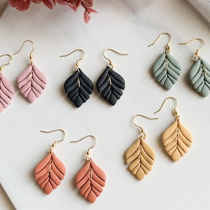 Pode incluir: Seis pares de brincos de folha pendurados em várias cores, incluindo rosa, preto, verde, laranja e bege. Cada brinco tem um gancho dourado e um design delicado de arame.