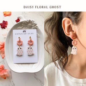 May include: A pair of pink and white ghost earrings with a daisy pattern. The earrings are made of polymer clay and have gold hooks. The earrings are displayed on a white card with the text "LOTUS OF CLAY" printed on it. The earrings are also shown on a model's ear.