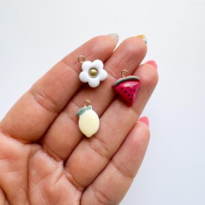May include: A collection of miniature charms featuring a white flower with a gold center, a lemon, and a watermelon slice. Each charm has a gold-colored loop for attaching to jewelry or other crafts. The charms are colorful and detailed.