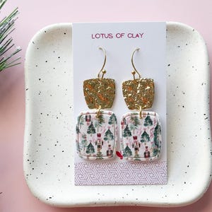 May include: Gold-toned, square earrings with a textured top and a patterned bottom. The bottom section features a festive design with Christmas trees and nutcracker figures. The earrings are displayed on a white tray with a pink background. The text "LOTUS OF CLAY" is visible.