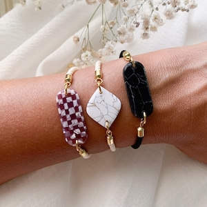 Op de afbeelding: Drie armbanden met verschillende gepatroneerde bedels. De bedels zijn rechthoekig en hebben een geruit patroon, een wit marmerpatroon en een zwart marmerpatroon. De armbanden zijn gemaakt van witte kralen en zwart touw.