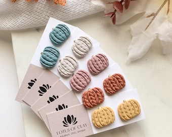 May include: Set of six pairs of polymer clay pumpkin earrings in various fall colors, including teal, beige, pink, orange, and yellow. Each pumpkin has a textured surface resembling a knitted sweater. The earrings are displayed on small cards with the text "LOTUS OF CLAY Modern, chic, unique".
