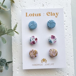 Op de afbeelding: Een set van zes handgemaakte polymer clay oorbellen. De oorbellen zijn in drie verschillende ontwerpen: een blauw geometrisch patroon, een roze bloemenpatroon en een wit en gouden patroon. De oorbellen worden getoond op een witte kaart met de tekst "Lotus Clay" en "Handmade Jewelry".