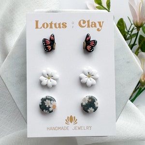 May include: A set of six handmade polymer clay earrings featuring a butterfly, a white flower, and a floral print. The earrings are displayed on a white card with the text "Lotus of Clay" and "Handmade Jewelry".