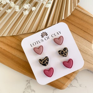 May include: A set of three heart-shaped earrings in pink and brown with a leopard print design. The earrings are displayed on a white card with the text "LOTUS OF CLAY".