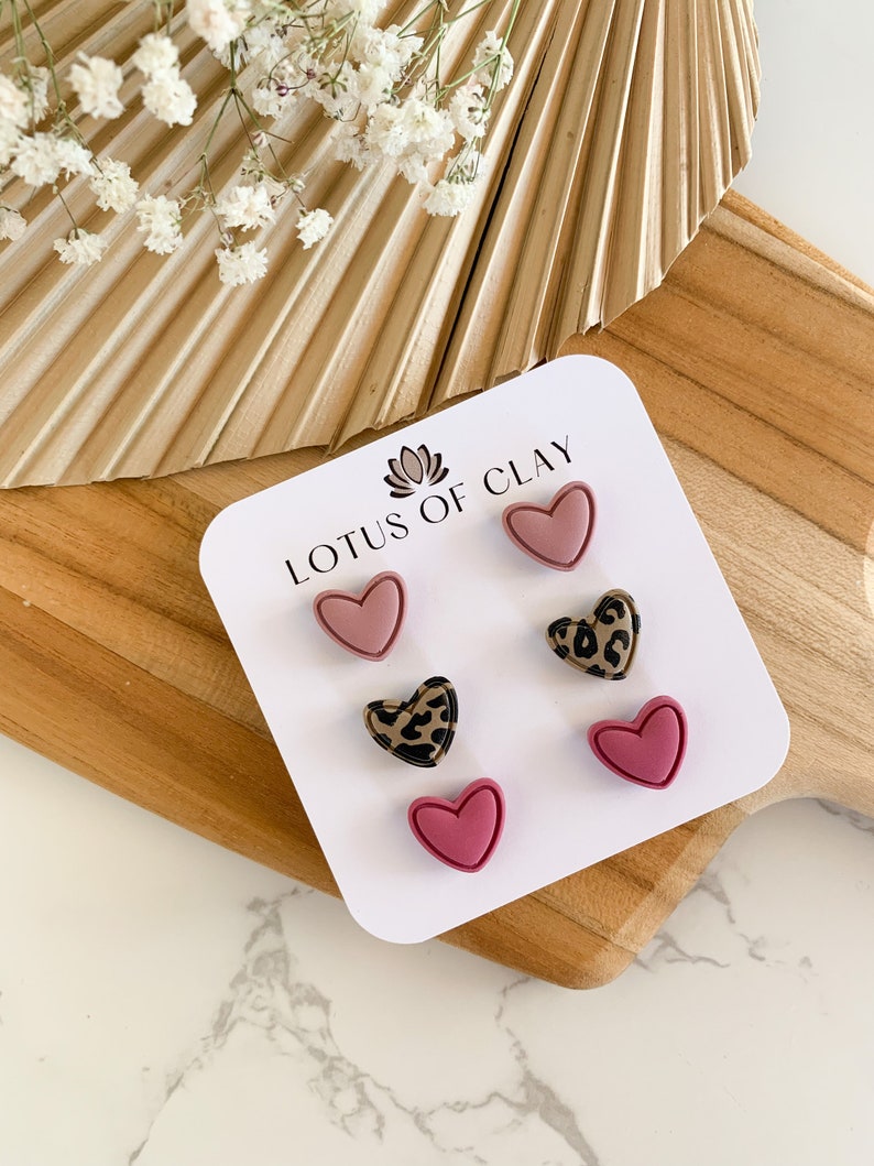 May include: A set of three heart-shaped earrings in pink and brown with a leopard print design. The earrings are displayed on a white card with the text "LOTUS OF CLAY".