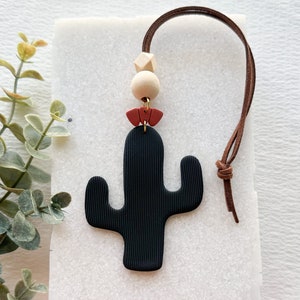 以下が含まれることがあります： 茶色の革紐が付いた黒と赤の サボテンの形をした車の芳香剤。サボテンは肋骨状の質感で、木製のビーズで飾られています。