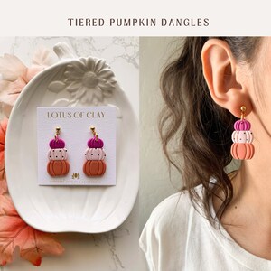 May include: A pair of tiered pumpkin dangle earrings with pink, white, and orange pumpkins. The earrings are handmade and feature a gold hook. The earrings are displayed on a white plate with a card that reads "LOTUS OF CLAY HANDMADE JEWELRY & ACCESSORIES".